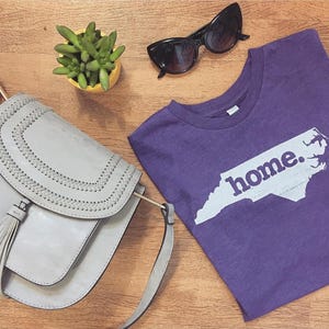 May include: A purple t-shirt with the word "home" and a white outline of the state of North Carolina. A gray purse with a tassel, black sunglasses, and a small yellow pot with a green plant are also visible.