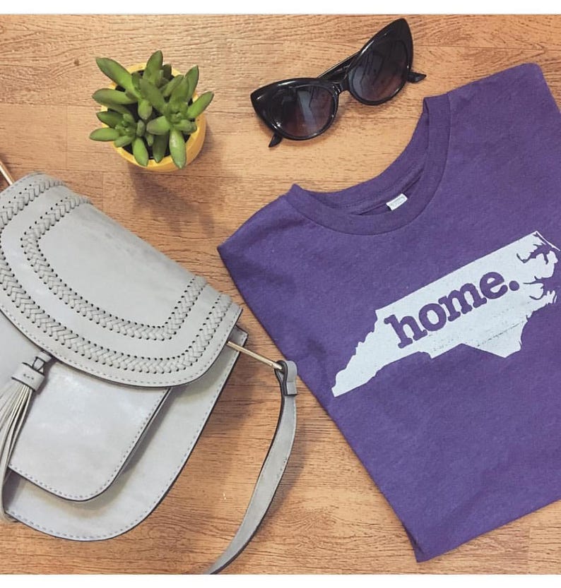 May include: A purple t-shirt with the word "home" and a white outline of the state of North Carolina. A gray purse with a tassel, black sunglasses, and a small yellow pot with a green plant are also visible.