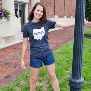 May include: A woman wearing a dark gray t-shirt with the word "home." printed inside the outline of the state of Ohio. She is also wearing blue denim shorts. The background includes a brick walkway and a building.