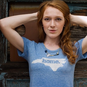 May include: A light blue t-shirt with the word "home" and a white outline of North Carolina. The person is wearing a necklace and has red hair. The background includes a brick wall and a wooden door.