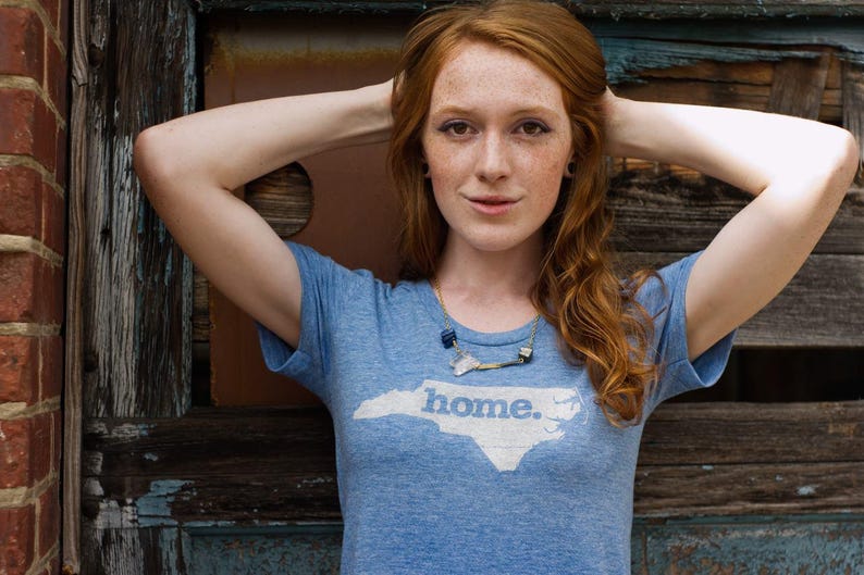 May include: A light blue t-shirt with the word "home" and a white outline of North Carolina. The person is wearing a necklace and has red hair. The background includes a brick wall and a wooden door.