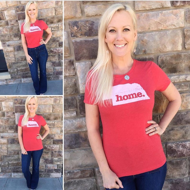 May include: A red short-sleeved t-shirt with the word "home" printed on it. The word "home" is printed in white on a white rectangle with a pink outline. The t-shirt is paired with blue jeans.