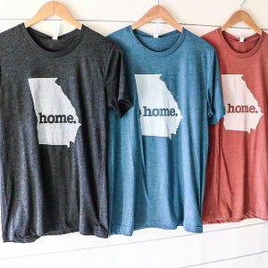 May include: Three short-sleeved t-shirts in charcoal, blue, and rust colors are displayed on wooden hangers. Each shirt features a white outline of the state of Georgia with the word "home." printed below it in a simple font.