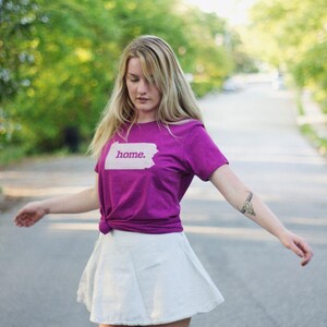 May include: A woman wearing a purple t-shirt with the word "home" printed on it, paired with a white skirt. The t-shirt is knotted at the waist. The background is a blurred street with trees.