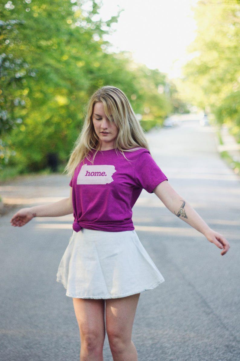 May include: A woman wearing a purple t-shirt with the word "home" printed on it, paired with a white skirt. The t-shirt is knotted at the waist. The background is a blurred street with trees.