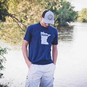 May include: A person wearing a navy blue t-shirt with the word "home" and a white outline of the state of Minnesota. The person is also wearing a gray and black baseball cap with a circular patch.