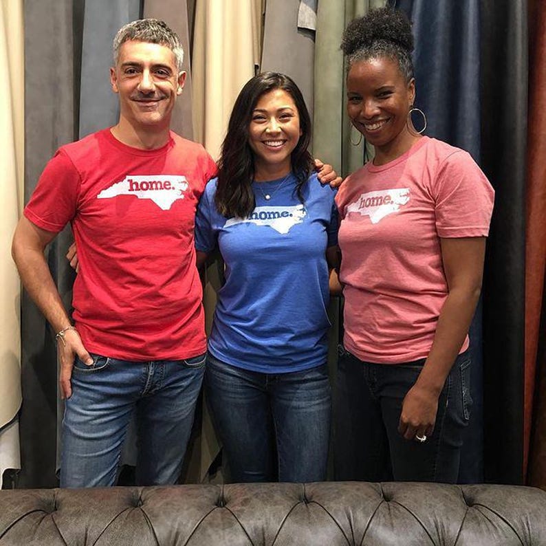 May include: Three people wearing t-shirts with the word "home" and a North Carolina outline. The shirts are red, blue, and pink. They are standing in front of a backdrop of fabric samples.
