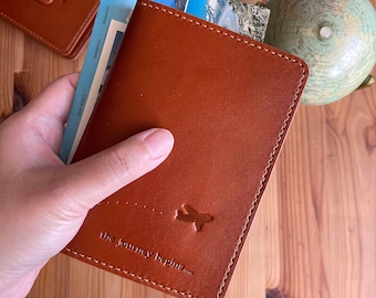 May include: A tan leather passport holder with a stitched border, holding postcards. The holder features an airplane graphic and the phrase "the journey begins...." on the front. Other leather travel accessories and a globe are in the background.