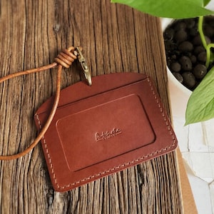 May include: A brown leather ID holder with a stitched border and a matching leather lanyard. The holder has a rectangular window and the word "Artchola Studio" embossed on the front. A gold-colored clasp secures the lanyard.