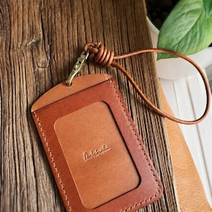 May include: A brown leather ID holder with a stitched border and a clear window. The holder has a matching leather lanyard and a gold-colored clasp. The text "Artchale Studio" is embossed on the front.