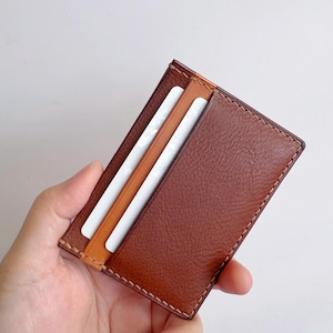May include: A brown leather card holder with a stitched border. The card holder has a light brown interior and contains two white cards. The leather has a textured appearance.