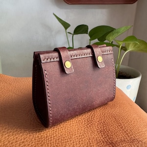 May include: A burgundy leather wallet with a flap closure and two brass-colored button clasps. The wallet is rectangular with visible stitching along the edges. The background includes a leather surface and a plant in a white pot.