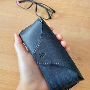 May include: A dark blue leather glasses case with a curved flap and white stitching. The case has a debossed logo and is next to a pair of black eyeglasses. The case is approximately 7 inches long.