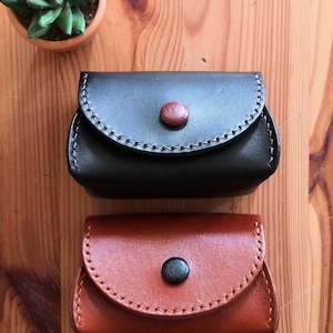 Könnte beinhalten: Zwei kleine, handgefertigte Leder-Münzbörsen. Die obere Börse ist dunkelbraun mit rotem Knopfverschluss und cremefarbenen Nähten. Die untere Börse ist hellbraun mit schwarzem Knopf und orangefarbenen Nähten. Im Hintergrund befindet sich eine kleine Topf-Sukkulente.