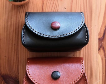 May include: Two small, handcrafted leather coin purses. The top purse is dark brown with a red button closure and cream stitching. The bottom purse is a lighter brown with a black button and orange stitching. A small potted succulent is in the background.