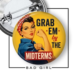 May include: A round pin-back button featuring a yellow background and a graphic illustration of a person with a red polka-dot headscarf and blue shirt, flexing an arm. The text reads GRAB EM by THE MIDTERMS in dark blue and red lettering.