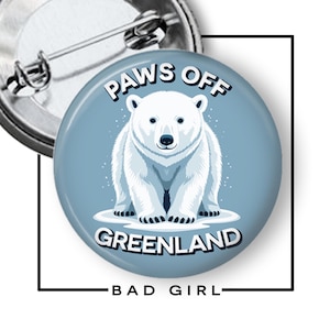 May include: A round, light blue button with a polar bear illustration. The text "PAWS OFF GREENLAND" arches above the bear, with "BAD GIRL" below. A silver pin back is visible.