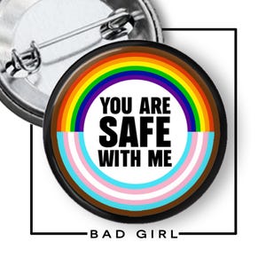 May include: A circular pin-back button featuring a white center with the black text 'YOU ARE SAFE WITH ME'. The design is surrounded by a rainbow arc on the top half and a light blue, pink, and white striped arc on the bottom half, with a black border.