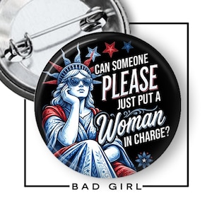 May include: A round pin-back button featuring a graphic of the Statue of Liberty wearing sunglasses. The design includes red, white, and blue colors with the text: CAN SOMEONE PLEASE JUST PUT A WOMAN IN CHARGE? The pin has a metal back with a pin closure.