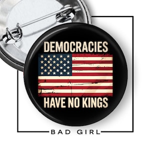 May include: A round pin-back button featuring a black background with the text DEMOCRACIES HAVE NO KINGS in a bold, light-colored font. Centered is a distressed-style American flag with red, white, and blue colors. The back shows a metal pin.