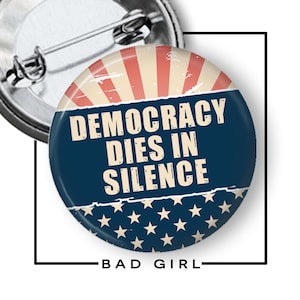 May include: A round button pin with a patriotic design. The top features red and beige stripes, transitioning to a dark blue field with white text that reads "DEMOCRACY DIES IN SILENCE." The bottom has white stars. The text "BAD GIRL" is below.