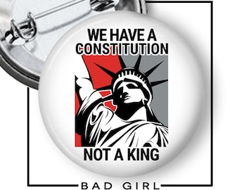 May include: A round pin-back button featuring a graphic of the Statue of Liberty in black and white against a red and grey background. The text WE HAVE A CONSTITUTION NOT A KING is printed in black capital letters above and below the illustration.