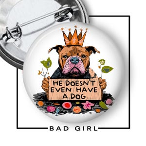 May include: A round button pin featuring a brown dog wearing a crown and holding a sign that reads "HE DOESN'T EVEN HAVE A DOG." The pin has a metal clasp on the back and the words "BAD GIRL" at the bottom.