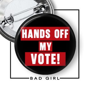 May include: A circular pin-back button featuring a black background with the white text "HANDS OFF MY VOTE!" displayed over three horizontal red rectangular bars. The back of the button shows a silver metal pin mechanism for attachment.