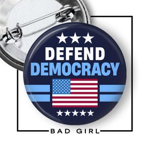 May include: A round, navy blue button with the words "DEFEND DEMOCRACY" in white and light blue. The button features the American flag and the text "BAD GIRL" at the bottom. The button has a pin back.