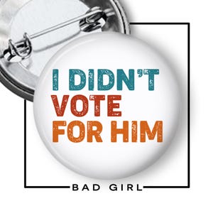 May include: A white button with the text "I DIDN'T VOTE FOR HIM" in teal, red, and orange. The button has a safety pin on the back and the words "BAD GIRL" at the bottom.