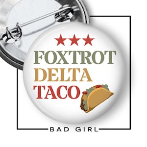 May include: A circular pin-back button featuring a white background with three red stars at the top. The text reads FOXTROT in olive green, DELTA in gold, and TACO in red. A graphic of a taco with brown meat and green lettuce is included.