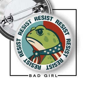 May include: A round button with a frog illustration wearing a patriotic scarf. The word "RESIST" is printed around the frog in a circular pattern. The text "BAD GIRL" is printed at the bottom.