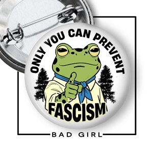May include: A round pin-back button featuring a green frog wearing a tan shirt and blue neckerchief, pointing forward. The text reads ONLY YOU CAN PREVENT FASCISM, surrounded by black pine tree silhouettes on a white background with a metal pin back.