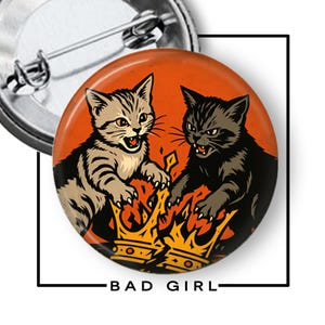 May include: A round pin-back button featuring an orange background with two kittens, one grey tabby and one black, facing each other with bared teeth above a broken yellow crown. The text BAD GIRL is printed in black below the circular design.