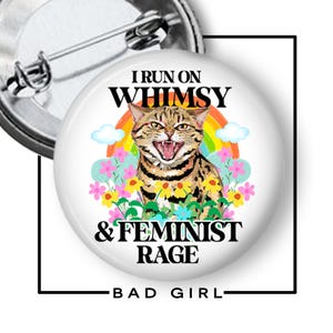 May include: A round pinback button featuring a brown tabby cat with an open mouth, surrounded by a colorful rainbow, clouds, and flowers. The text reads I RUN ON WHIMSY & FEMINIST RAGE. The back of the pin shows a silver metal pin mechanism.