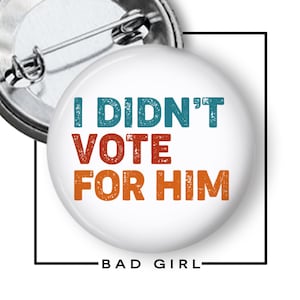 May include: A round white pin-back button featuring the text I DIDN'T VOTE FOR HIM in a distressed font. The words I DIDN'T VOTE are in a teal blue color, while FOR HIM is in a burnt orange color. The back of the button shows a metal pin mechanism.