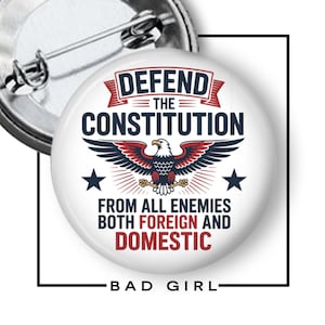 May include: A round, white button with the text "DEFEND THE CONSTITUTION" and an eagle graphic. The button also includes the text "FROM ALL ENEMIES BOTH FOREIGN AND DOMESTIC" and "BAD GIRL". The button has a safety pin backing.