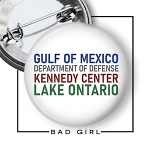 May include: A round white pin-back button featuring four lines of text in different colors: GULF OF MEXICO in blue, DEPARTMENT OF DEFENSE in gray, KENNEDY CENTER in maroon, and LAKE ONTARIO in green. The word BAD GIRL is printed below the button.