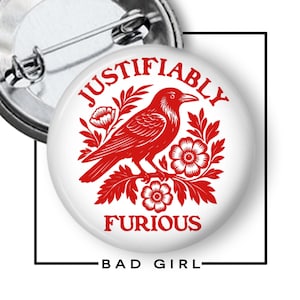 May include: A white button pin with a red illustration of a crow perched on flowers, with the words "JUSTIFIABLY FURIOUS" and "BAD GIRL" in red text. The pin has a metal clasp.