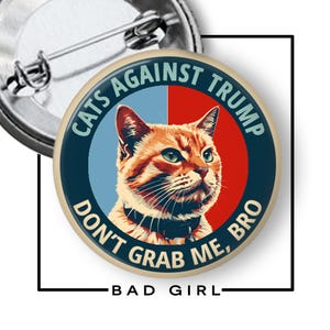 May include: A round pin-back button featuring a ginger cat centered against a split light blue and red background. The text CATS AGAINST TRUMP is arched at the top, and DON'T GRAB ME, BRO is arched at the bottom in dark blue lettering.