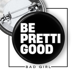 May include: A black button with the words "BE PRETTI GOOD" in bold white letters. The phrase "BAD GIRL" is written in white at the bottom. A silver pin backing is visible.
