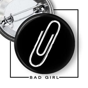 May include: A round pin-back button featuring a black background with a white, hand-drawn style paperclip graphic. The text "BAD GIRL" is printed in black capital letters below the button, which has a metal pin mechanism on the back.