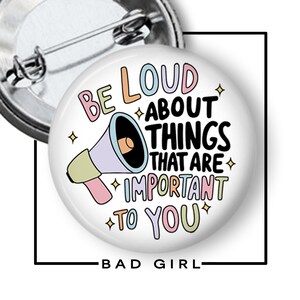 May include: A round pin-back button featuring a pastel-colored megaphone illustration and the text BE LOUD ABOUT THINGS THAT ARE IMPORTANT TO YOU. The design includes small star accents in black, pink, blue, and yellow on a white background.