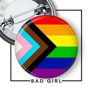 May include: A round button pin with a rainbow Pride flag design, including the transgender flag and a black and brown chevron. The text "BAD GIRL" is printed in black at the bottom of the image. The pin has a metal backing.