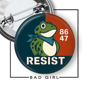 May include: A round button with a cartoon frog wearing a blue bandana. The button is split into two sections, one navy blue and one rust-colored. The word "RESIST" is printed in white, with the numbers "86 47" above it. The text "BAD GIRL" is below the button.