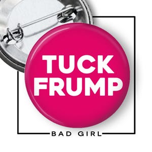 May include: A circular pin-back button featuring a vibrant hot pink background with the bold white text TUCK FRUMP centered on the front. The back of the button is metallic silver with a standard pin mechanism for attachment to clothing or fabric.