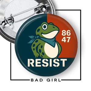 May include: A circular pin-back button featuring a green frog wearing a blue neckerchief. The background is split between dark teal and rust orange. The text "RESIST" appears in white at the bottom, with the numbers "86 47" printed in white on the right.