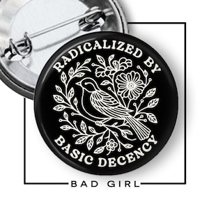 May include: A black button pin with white text and a bird and floral design. The text reads "RADICALIZED BY BASIC DECENCY" and "BAD GIRL". The pin has a metal clasp on the back.