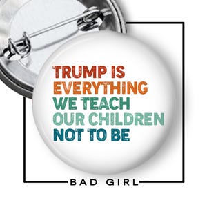 May include: A white button pin with the text "TRUMP IS EVERYTHING WE TEACH OUR CHILDREN NOT TO BE" in orange, green, and blue. The pin has a metal clasp and the words "BAD GIRL" at the bottom.