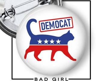 May include: A white button pin with a cat silhouette in red, blue, and white, with the word "DEMOCAT" in a red rectangle. The pin also features the text "BAD GIRL" in black.